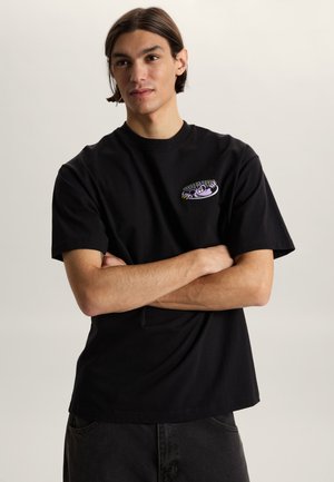 Joven con cabello hasta los hombros viste una camiseta negra con un pequeño diseño de araña morada en el pecho, brazos cruzados, fondo neutro.
