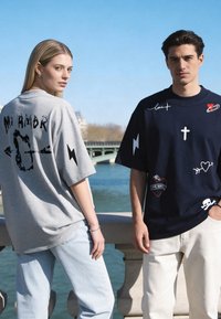 Jeune femme et homme portant des t-shirts graphiques oversize avec des motifs sur le thème de l'amour, debout près d'une rambarde au bord d'une rivière sous un ciel bleu.