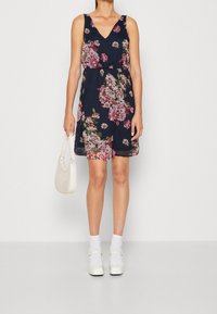 Blommönstrad marinblå klänning med V-ringning, ärmlös design, cinchad midja och lätt tyg. Matchad med en vit väska och skor.