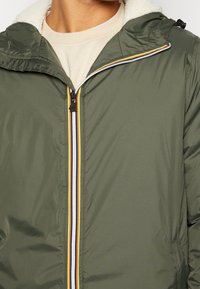 Giacca verde oliva con cappuccio, con un colletto interno bianco e soffice, dettagli della zip multicolore e un esterno leggero e liscio.