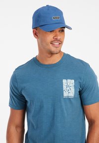 Blauwe pet met een logo-opdruk aan de voorkant, gecombineerd met een blauw T-shirt met korte mouwen dat een witte, abstracte print op de borstzak heeft.