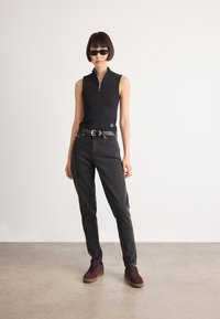 Kvinne som står i en svart ermeløs glidelås-topp, mørkegrå jeans med svart belte, lilla joggesko og svarte solbriller mot en hvit vegg.