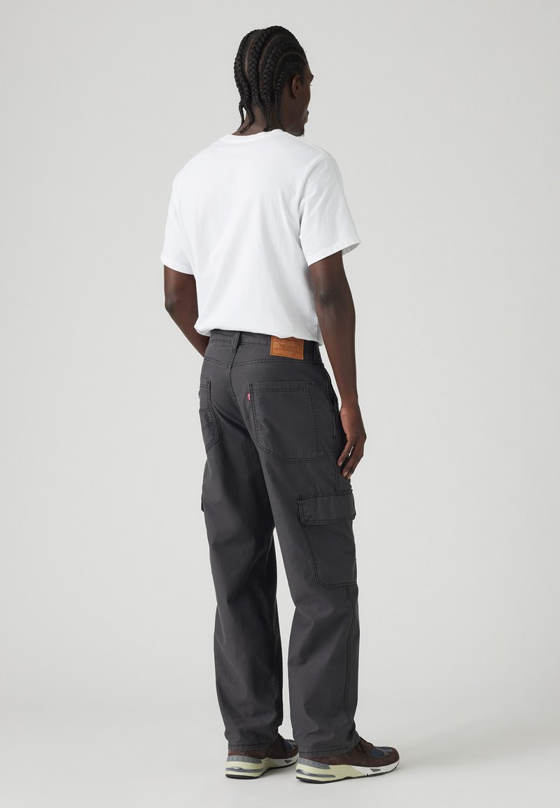 Pantalones cargo de color carbón con múltiples bolsillos, costuras reforzadas y un parche de cuero marrón en la parte trasera. Combinados con una camiseta blanca.