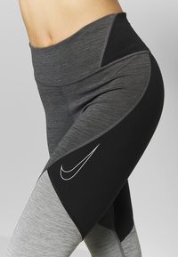 Legginsy z wysokim stanem, w połączonym ciemnoszarym i czarnym materiale, z białym logo Nike, gładką teksturą i sportowym krojem.