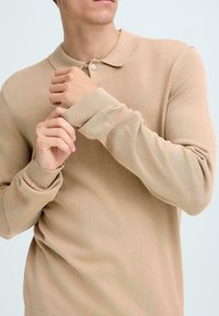Beige strikket piké skjorte med klassisk krage, ribbede mansjetter og myk tekstur, båret av en person som justerer ermene.