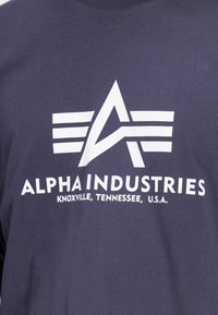 Chemise bleue avec logo blanc Alpha Industries, un « A » stylisé avec trois lignes horizontales, et le texte « Knoxville, Tennessee, U.S.A. » en dessous.