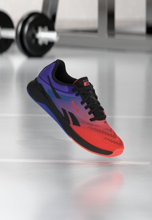 Zapatilla de correr en colores púrpura, rojo y negro con cordones negros, flotando sobre un suelo de gimnasio reflectante con mancuernas desenfocadas en el fondo.