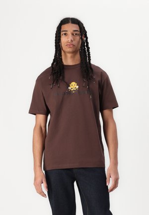 Brown cotton t-shirt featuring a yellow cartoon monkey graphic and text in black, short sleeves, and a relaxed fit. Denim jeans complete the look.