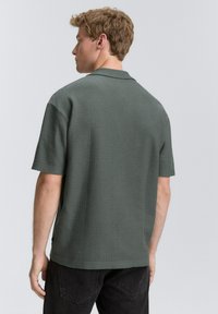 Ung man med kort lockigt hår som bär en strukturerad, mörkgrön kortärmad polo och svarta byxor, vänd bort från kameran mot en neutral bakgrund.