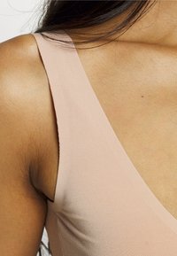 Jasnoróżowy, bezrękawnik z głębokim dekoltem w kształcie litery V, prążkowaną teksturą i gładkim materiałem, podkreślający ramiona i okolice obojczyków.