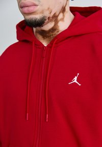 Haut zippé rouge en tissu doux, arborant un logo Jumpman blanc sur la poitrine, avec des cordons de serrage réglables et une fermeture éclair à l'avant.