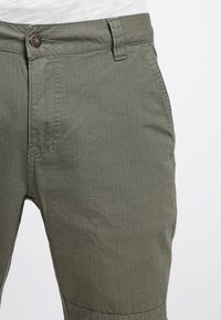 Närbild av olivgröna byxor med textilstruktur, synlig knapp, bältesöglor och en vit skjorta instoppad.