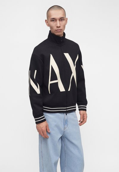Homme portant une veste noire zippée avec de grandes lettres blanches « AX » et des poignets rayés, associée à un jean ample bleu clair.