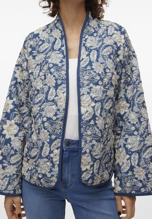 Femme portant une veste matelassée bleue avec des motifs floraux crème, associée à un jean bleu et une chemise blanche.
