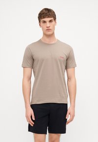 T-shirt en coton beige à manches courtes, avec un col ras du cou et un petit logo rouge "HUGO" sur la poitrine gauche. Associé à un short noir.