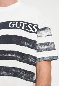 Λευκό T-shirt με ναυτικές οριζόντιες ρίγες και τη λέξη "GUESS" σε έντονα λευκά γράμματα στο στήθος. Μαλακή βαμβακερή υφή.