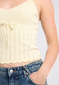 Cream knit camisole with scalloped edges and a textured pattern, featuring a tie at the waist. Paired with distressed denim jeans.