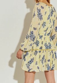 Vestido floral amarillo con patrones azules, mangas largas abullonadas, cintura ceñida y dobladillo con volantes y detalles de encaje.