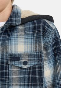Camicia a scacchi blu con cappuccio foderato in pile; presenta tonalità di blu scuro e chiaro, chiusura a bottoni e una tasca sul petto con etichetta.