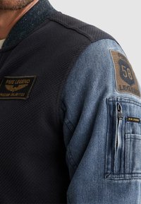Primer plano de una chaqueta con tela negra texturizada y manga de denim azul con parches de PME Legend y un bolsillo con cremallera.