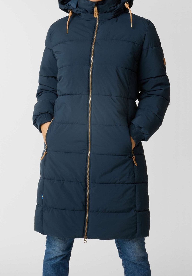 Doudoune bleu marine avec capuche, dotée d'une fermeture éclair à l'avant, de poignets côtelés et de deux poches latérales avec des accents beige. Design matelassé.