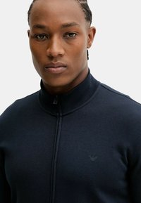 Primo piano di un uomo con capelli intrecciati che indossa un maglione blu navy con zip e un piccolo logo ricamato sul petto, su uno sfondo neutro.