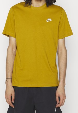 Person iført guldfarvet Nike T-shirt med hvidt logo på brystet og sorte shorts, stående med armene afslappet langs siden.