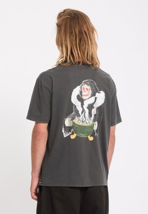 Personne aux cheveux blonds mi-longs porte un t-shirt gris foncé avec un dessin représentant une silhouette à capuche tenant un bol vert rempli de cristaux blancs.