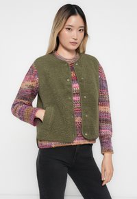 Groene fleecvest met een ronde kraag en drukknoopsluiting, gedragen over een multicolor gebreid sweater. Het vest heeft twee zijzakken.