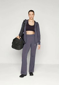 Sporta apģērbs, kas ietver tumši pelēku ar rāvējslēdzēju aizveramu džemperi, melnu īsu topu un atbilstošus augsta jostasvietas bikses, kopā ar melnu apavu un sporta somu.