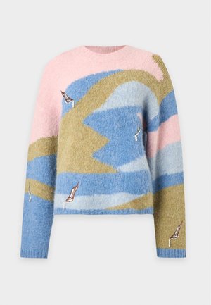 Femme portant un pull pastel à motif abstrait, une longue jupe blanche avec des détails œillets et des ballerines marron à lacets, debout sur un fond uni.