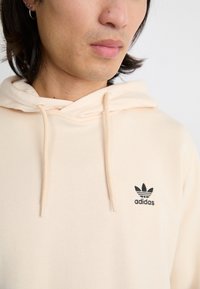 Felpa beige realizzata in morbido tessuto, con tasca frontale, cappuccio con lacci e un logo Adidas ricamato in nero sul lato sinistro del petto.
