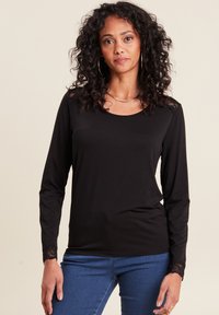 Schwarzes langärmliges Oberteil aus weichem Stoff, mit rundem Halsausschnitt und Spitzenakzenten an den Ärmeln. Kombiniert mit blauen Jeans.