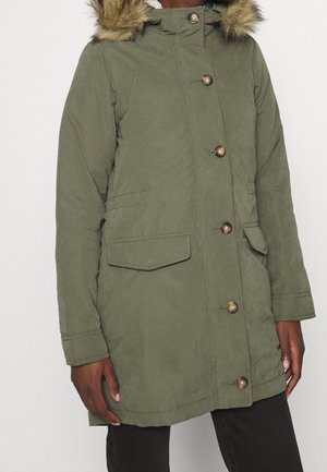 Femme portant un manteau d'hiver vert olive avec capuche bordée de fourrure et grands boutons, avec poches à rabat et manches longues.