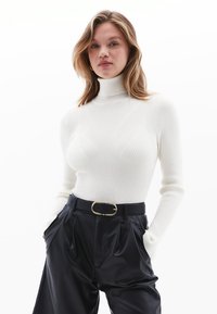 Weißer gerippter Rollkragenpullover mit langen Ärmeln, kombiniert mit hochgeschnittenen dunklen Hosen und einem schwarzen Gürtel mit goldener Schnalle.