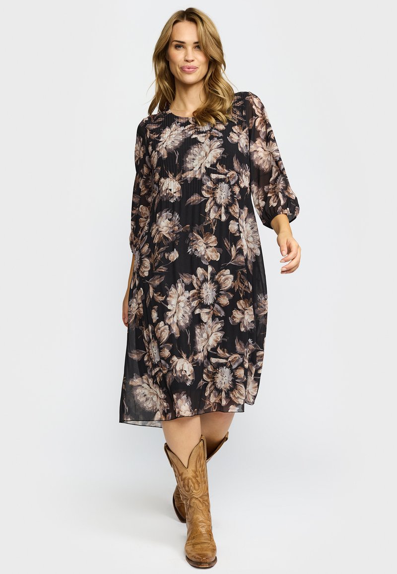 Kvinne som går i en svart blomstrete midi-kjole med trekvartlange ermer og brune cowboystøvler mot en ensfarget bakgrunn.