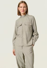 Lichtgrijs langemouw shirt met een hoge hals, twee frontzakken en gouden knopenaccenten op de schouders. Getailleerde pasvorm met een soepele textuur.