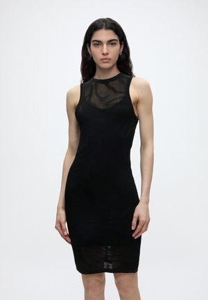 Ženska z dolgimi temnimi lasmi, oblečena v oprijeto črno obleko brez rokavov z teksturiranim vzorcem, stoji pred enobarvnim belim ozadjem.