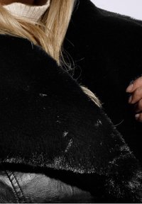 Schwarzer Kunstpelzkragen mit weicher, glatter Textur, über einer schwarzen Lederjacke geschichtet. Zeigt eine Nahaufnahme des Kragens und der Hand.