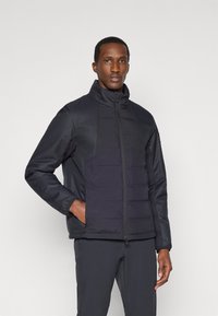 Casaco preto acolchoado com gola alta e fecho éclair na frente. Apresenta painéis contrastantes, punhos elásticos e bolsos laterais. Materiais lisos e texturizados.