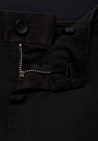 Pantaloni in tessuto nero con chiusura a bottone e cerniera metallica. Presentano una texture liscia, dettagli di cucitura e un piccolo anello in tessuto vicino alla cerniera.