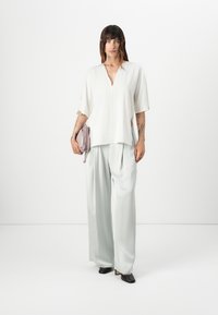 Loose-fitting white blouse with a V-neck, short sleeves, paired with light gray wide-leg trousers. Holds a small lilac handbag. Black shoes.
