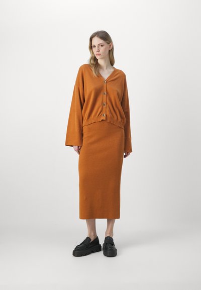Un pull en tricot orange avec des boutons et des manches larges, associé à une jupe longue assortie. Des chaussures à plateforme noires complètent la tenue.