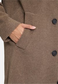 Cappotto di lana marrone con una texture morbida, dotato di tasca laterale e bottoni neri. Il design include rever a scollo a V e una silhouette sartoriale.