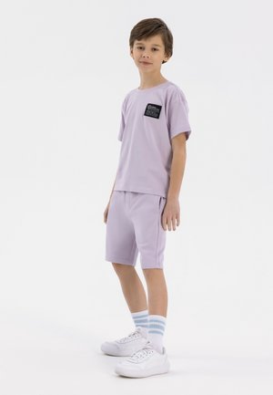 T-shirt en coton lilas à manches courtes avec une poche, assortie à un short. Des baskets blanches et des chaussettes rayées bleu et blanc complètent le look.