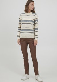 Helle Cremefarbene Pullover mit horizontalen blauen, braunen und orangefarbenen Streifen. Kombiniert mit braunen, figurbetonten Hosen und weißen Sneakers.