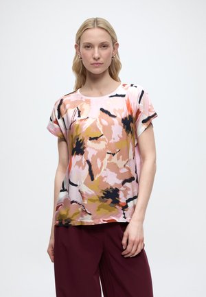 Kobieta o długich blond włosach, ubrana w krótki rękaw koszuli z abstrakcyjnym kwiatowym wzorem w kolorach różowym, pomarańczowym i brązowym, w połączeniu z ciemnoczerwonymi spodniami.