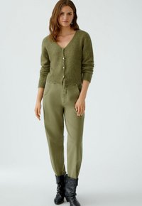 Olivgrüner Wolle-Mix Cardigan mit V-Ausschnitt, Knopfverschluss und geripptem Saum. Kombiniert mit hellgrünen, weiten Hosen und schwarzen Stiefeln.