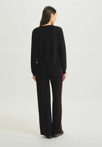 Pull noir à coupe décontractée avec un col rond, associé à un pantalon large noir avec des rayures rouges sur les côtés. Texture en maille lisse et design confortable.