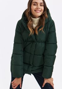 Zielona kurtka puchowa z kapturem, z pikowanym wzorem, teksturowanym materiałem i regulowanym sznurkiem. Nosi na beżowym golfie.